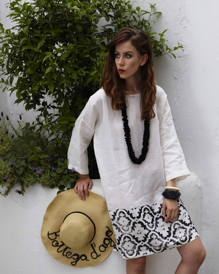 Look estivo minimal: modella con abito in lino bianco e nero, collana maxi e cappello di paglia "Bottega d’ago". Outfit casual elegante, scatto di moda mediterranea.