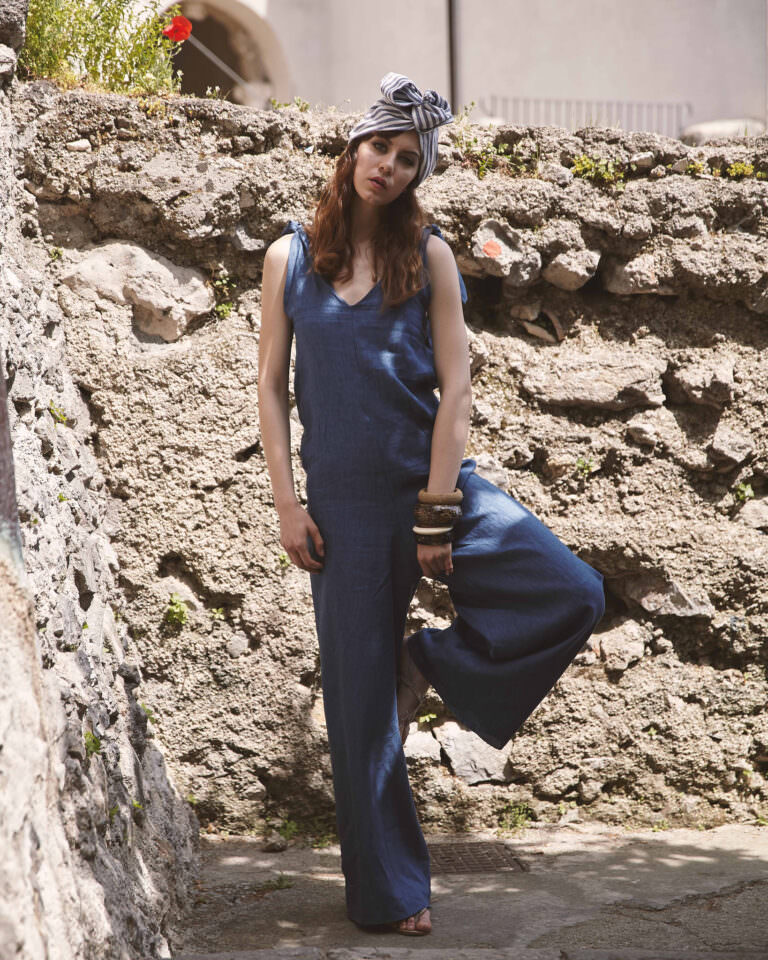 Donna con tuta blu in lino e turbante a righe, posa contro un muro di pietra – Shooting moda estiva by Bulb Studio