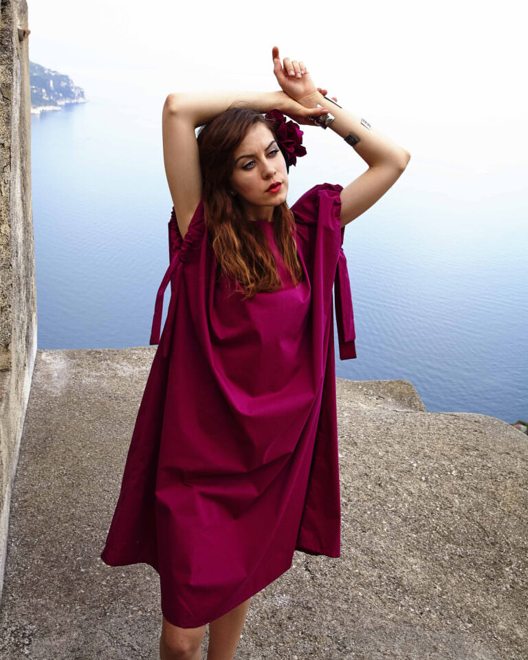 Modella con abito fucsia su una scogliera con vista sul Costiera Amalfitana mare, moda estiva – Fotografia editoriale di Bulb Studio Fashion
