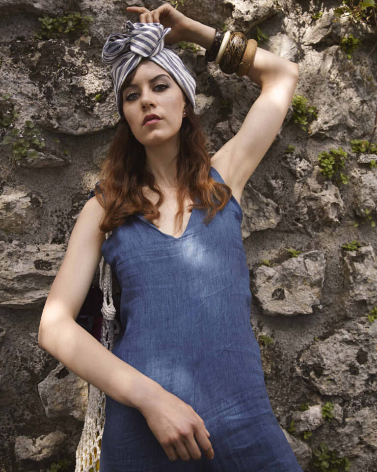 Modella con abito in denim leggero e turbante a righe bianche e blu, accessori boho in legno e metallo. Shooting moda estate su sfondo rustico di pietra